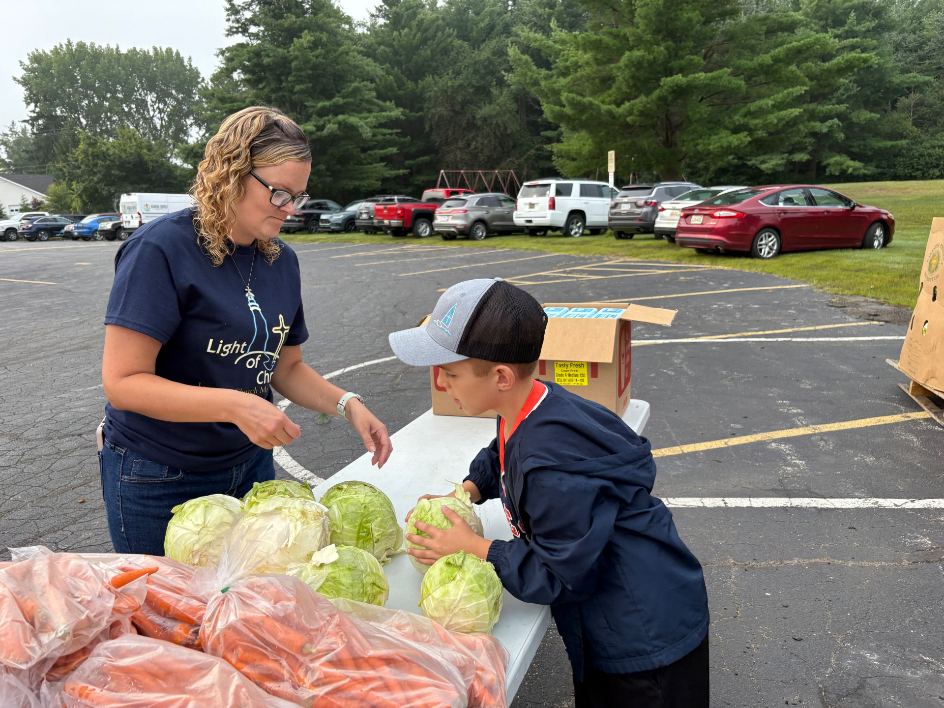 A volunteer and boy stand at a folding table covered with cabbage and bags of carrots.